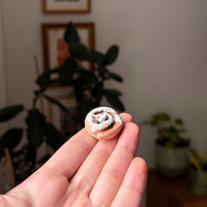 May include: A small, detailed cinnamon roll sculpture with a light brown base and white icing. The miniature food art is held in a hand, highlighting its size and craftsmanship. A unique decorative item, reminiscent of a sweet treat.