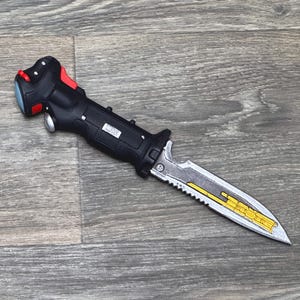 Puede incluir: Un cuchillo negro y plateado con una hoja dentada y un mango negro con un botón rojo. El cuchillo tiene una franja amarilla que recorre la hoja.