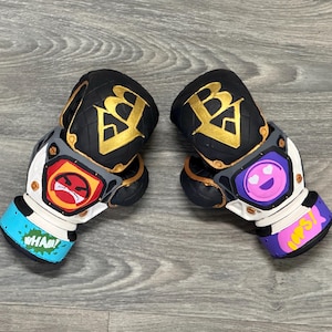 Puede incluir: Par de guantes de boxeo personalizados. Los guantes son negros con detalles dorados y presentan un logotipo "B" estilizado. Un guante tiene un gráfico de cara enojada, el otro una cara sonriente. Los puños tienen las palabras "WHAM!" y "OOPS!"