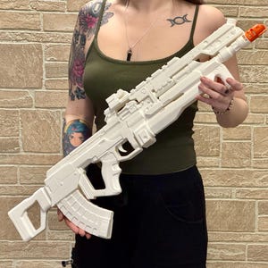 May include: A large, off-white toy rifle with an orange tip. The rifle features a complex design with multiple components, including a magazine, scope, and various attachments. The toy gun is held against a brick wall background.
