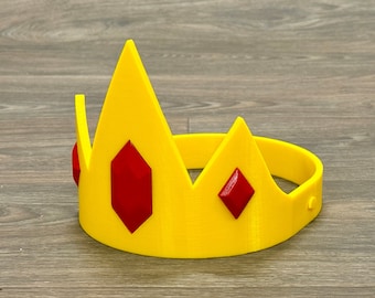 May include: A yellow plastic crown with three red gem accents. The crown is designed to be worn as a headband.