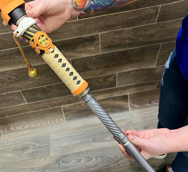 May include: A decorative, wooden-handled sword with a black and silver metal blade. The handle is wrapped in a beige fabric with a gold and black pattern. The sword is held by a person's hand.