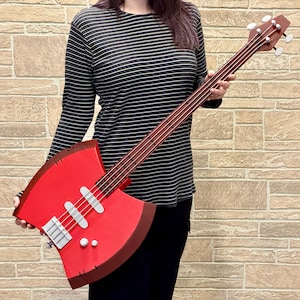 Puede incluir: Un bajo rojo con un cuerpo en forma de hacha. El instrumento tiene un mástil y clavijero marrones, trastes plateados y clavijas blancas. El cuerpo es de un rojo vibrante con ribetes marrones, y el diseño recuerda a un hacha de batalla.