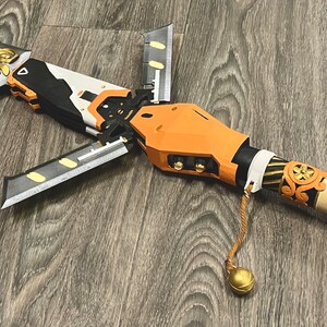 May include: A detailed, multi-part, orange and black prop weapon with a gold and black handle and a gold tassel. The weapon has a futuristic design and appears to be made of plastic.