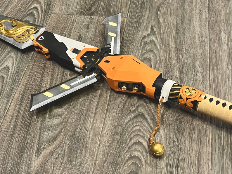 May include: A detailed, multi-part, orange and black prop weapon with a gold and black handle and a gold tassel. The weapon has a futuristic design and appears to be made of plastic.