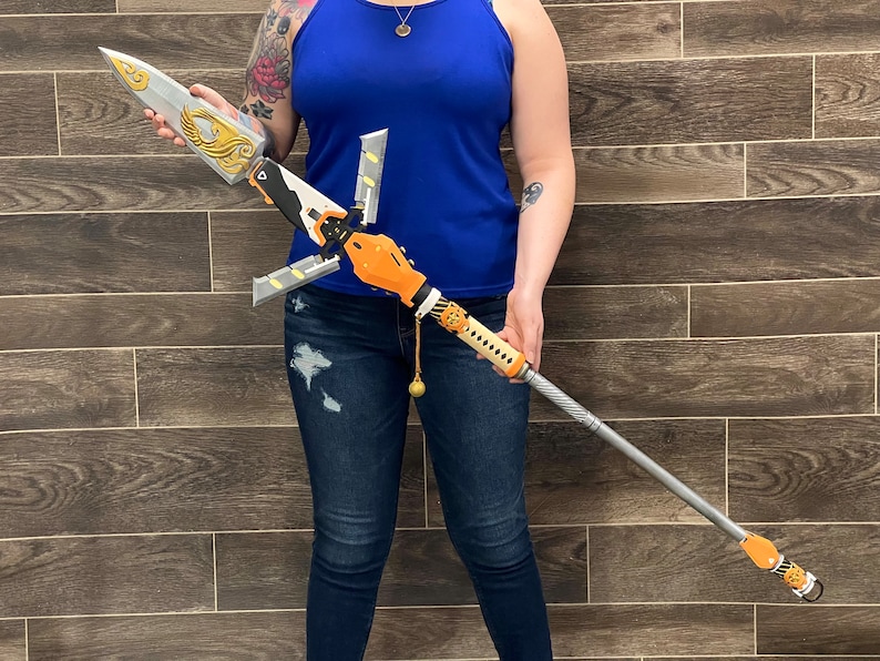 May include: A silver and orange spear with a decorative design. The spear has a long, thin shaft and a sharp point. The spear is held by a person wearing blue jeans.