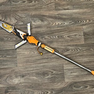 May include: A silver and gold spear with an orange handle and a decorative design. The spear is lying on a wooden surface.