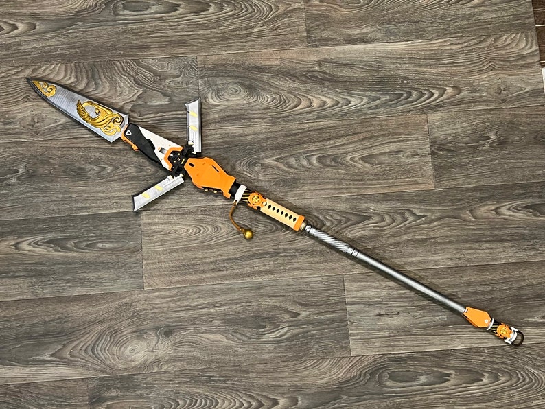 May include: A silver and gold spear with an orange handle and a decorative design. The spear is lying on a wooden surface.