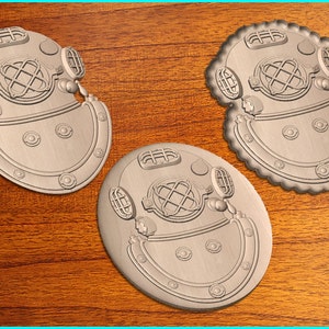 May include: Three silver-coloured metal diving helmet ornaments. The ornaments are round and have a raised design of a diving helmet with a window and various details. The ornaments are on a wooden surface.