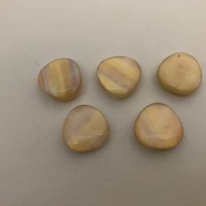 May include: Five irregular, rounded buttons with a smooth, iridescent finish. Each button displays a soft, creamy beige colour with subtle pearlescent highlights that shift in the light, creating a polished, reflective surface on each piece.