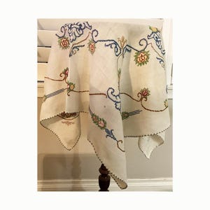 May include: A square off-white linen tablecloth featuring intricate cross-stitch embroidery in blue, green, pink, and brown. The design includes scrolling floral motifs and decorative borders with a scalloped edge finished in dark brown thread.