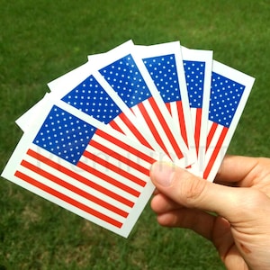 Puede incluir: Un conjunto de seis pequeños tatuajes temporales con la bandera de Estados Unidos. Los tatuajes son blancos con estrellas y rayas rojas y azules.