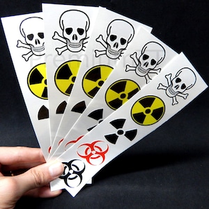 Może przedstawiać: Zestaw sześciu tymczasowych tatuaży z czarno-białymi motywami czaszek i skrzyżowanych kości, symbolami zagrożenia biologicznego i promieniotwórczości. Tatuaże są wydrukowane na białym papierze.