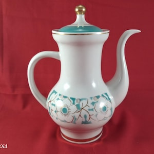 Puede incluir: Cafetera de cerámica blanca con un diseño floral turquesa y dorado. La cafetera tiene un asa y un pico curvos, una tapa turquesa y un adorno dorado. La base tiene una banda dorada. El fondo es rojo.