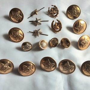 May include: A collection of vintage brass buttons and insignia. The buttons are round with a star design in the center. Some buttons are oval-shaped. The insignia includes star pins and wreath-shaped pieces. The items are in various sizes and have a gold finish.