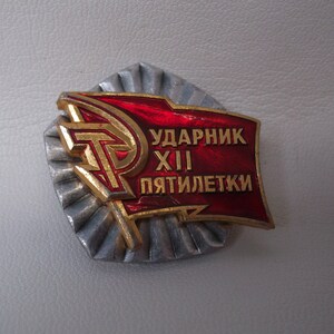 May include: A red and gold metal badge with the text "Ударник XIII пятилетки" which translates to "Shock Worker of the 13th Five-Year Plan".