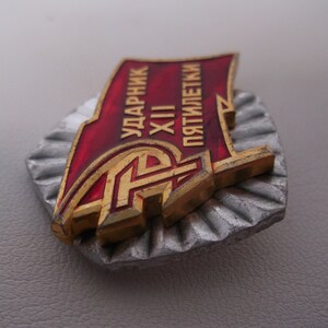 May include: A red and gold metal badge with the text "УДАРНИК XII ПЯТИЛЕТКИ" (meaning "Shock Worker of the 12th Five-Year Plan").