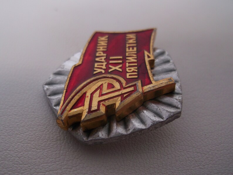 May include: A red and gold metal badge with the text "УДАРНИК XII ПЯТИЛЕТКИ" (meaning "Shock Worker of the 12th Five-Year Plan").