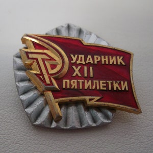 May include: A red and gold metal pin with a hammer and sickle design and the text "Ударник XII Пятилетки" which translates to "Shock Worker of the 12th Five-Year Plan".