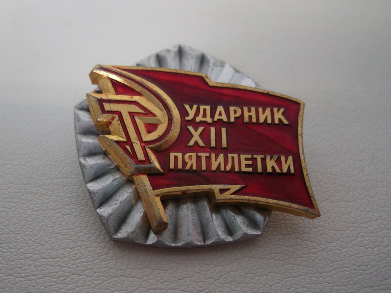 May include: A red and gold metal pin with a hammer and sickle design and the text "Ударник XII Пятилетки" which translates to "Shock Worker of the 12th Five-Year Plan".
