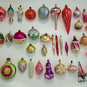 May include: A collection of assorted vintage glass Christmas ornaments in various shapes, sizes, and colors including red, green, gold, silver, and pink. Some feature metallic finishes, glitter, or patterns. One ornament is a clock face with hands.