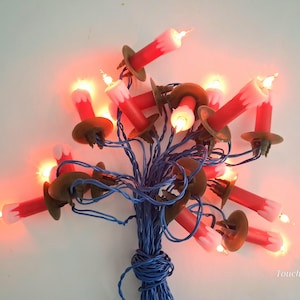 Op de afbeelding: Een snoer met elektrische kerstverlichting bestaande uit rode kaarsvormige lampjes met witte druppeldetails. Elk lampje zit in een bruine kunststof voet en is verbonden met blauwe bedrading. De lampjes branden en geven een warm licht.