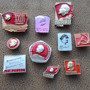 May include: A collection of ten vintage enamel pins featuring portraits of Lenin, a hammer and sickle, and other Soviet symbols. The pins are red, white, and gold in color and have various text in Cyrillic.