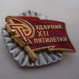 May include: A red and gold metal pin with the text "Ударник XII Пятилетки" (meaning "Shock Worker of the 12th Five-Year Plan").