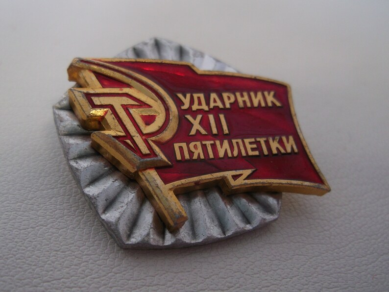May include: A red and gold metal pin with the text "Ударник XII Пятилетки" (meaning "Shock Worker of the 12th Five-Year Plan").