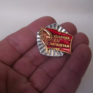 May include: A red and gold metal pin with the text "Ударник XII пятилетки" which translates to "Shock Worker of the 12th Five-Year Plan".