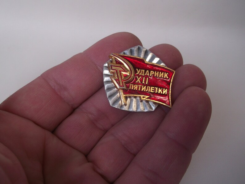 May include: A red and gold metal pin with the text "Ударник XII пятилетки" which translates to "Shock Worker of the 12th Five-Year Plan".