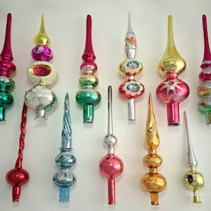 Op de afbeelding: Een collectie van elf vintage glazen kerstboompieken in diverse vormen en kleuren, waaronder rood, goud, groen, blauw en zilver. Elke piek heeft een puntige vorm, bolvormige secties en reflecterende metallic afwerkingen.