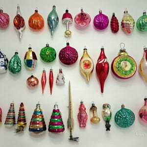 Puede incluir: Una colección de adornos navideños de vidrio vintage surtidos en varias formas, incluyendo esferas, gotas, remates y diseños figurativos como una casa, un búho y un cohete. Los adornos presentan colores metálicos vibrantes, purpurina y detalles.