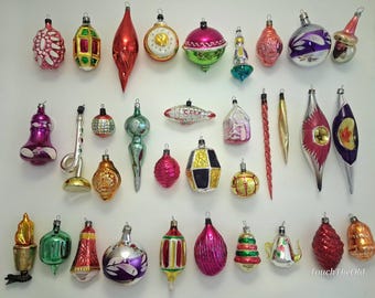 May include: A collection of thirty-five vintage glass Christmas ornaments in various shapes, including spheres, finials, and figurals. The ornaments feature diverse colors such as red, green, gold, silver, pink, and purple, with some displaying painted details.