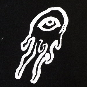 May include: A rectangular black fabric patch featuring a white screen-printed graphic of a single eye centered within an oval shape, with three distinct, fluid, dripping lines extending downward from the bottom of the oval form.