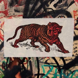 Pode incluir: Um patch de tecido retangular com uma ilustração detalhada de um tigre-dentes-de-sabre em tons de laranja e vermelho. O tigre é retratado em uma pose de caminhada com a boca aberta. O tecido tem uma borda crua.
