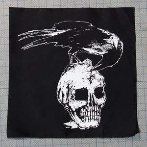 Könnte beinhalten: Ein schwarz-weißes Patch mit einem Raben, der auf einem Schädel sitzt. Der Rabe blickt nach rechts, der Schädel nach vorne. Das Patch ist aus schwarzem Stoff mit weißem Design.