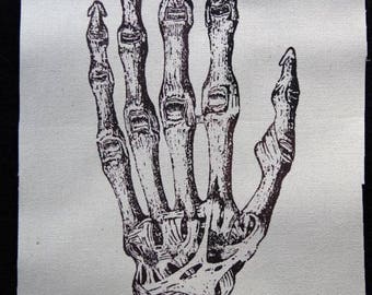 May include: A rectangular fabric patch featuring a detailed black ink illustration of a human hand skeleton. The drawing highlights the intricate bone structure, joints, and ligaments of the hand and wrist against a plain off-white background.