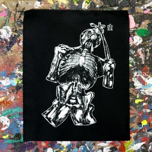 Op de afbeelding: Zwart vierkantige patch met een witte illustratie van een skelet dat een touw vasthoudt. Het skelet is afgebeeld op een gestileerde, artistieke manier. Een klein symbool staat boven het hoofd van het skelet. De achtergrond is een kleurrijk, met verf bespat oppervlak.