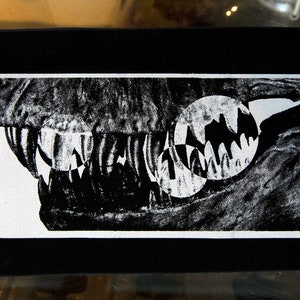May include: Black and white image of a large, toothy mouth with a white background. The mouth is open wide, revealing sharp teeth and a dark interior. The image is framed in a black border.