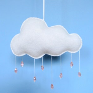 May include: A white felt cloud mobile with pink crystal raindrops. The cloud hangs from a white string against a light blue backdrop. The teardrop-shaped raindrops are a delicate pink and sparkle.