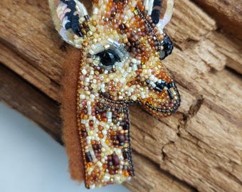 May include: A handcrafted giraffe brooch, meticulously beaded with a variety of brown, tan, and black beads. The giraffe's head and neck are detailed with a spotted pattern, and it has small black ears and tufts of black hair. The brooch is set against a wooden background.
