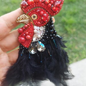 Pode incluir: Broche artesanal em forma de galo. O galo apresenta uma crista e barbela de contas vermelhas, um bico dourado e um corpo adornado com contas pretas e penas pretas. Cristais e pérolas brilhantes adicionam detalhes.