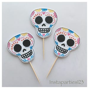May include: Three white sugar skull cupcake toppers with colourful floral designs and black eyes. The toppers are on wooden sticks.