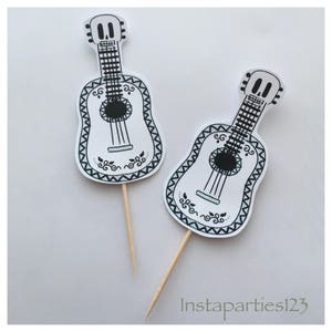 May include: Two white guitar cupcake toppers with black details and a skull design. The toppers are on wooden sticks.