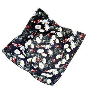 May include: A black fabric bowl cozy with a white daisy and ladybug pattern. The cozy is square-shaped and has a quilted appearance. The design features white daisies with yellow centers and red ladybugs scattered across the surface.