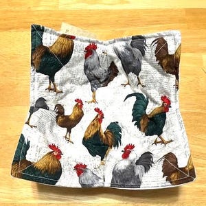 May include: A white fabric pot holder featuring a repeating pattern of colorful roosters. The roosters are depicted in various poses with red combs, brown, black, and gray feathers. The background has a subtle newspaper print.