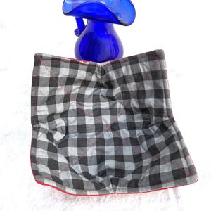 May include: A blue glass pitcher with a ruffled rim sits above a black, white, and gray plaid bowl cozy with a red trim. The cozy is square and quilted.