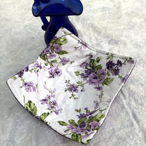 May include: A white fabric bowl cover with a floral pattern of purple flowers and green leaves. The bowl cover is square-shaped with a dark purple trim. A cobalt blue glass vase is in the background.