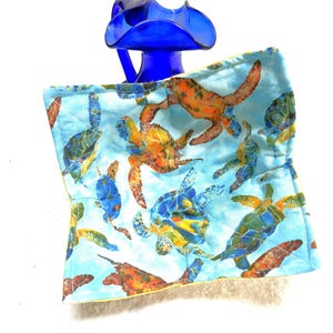 May include: A blue quilted pot holder with a sea turtle pattern. The turtles are in shades of blue, orange, and green against a light blue background. A cobalt blue glass bottle is in the background.